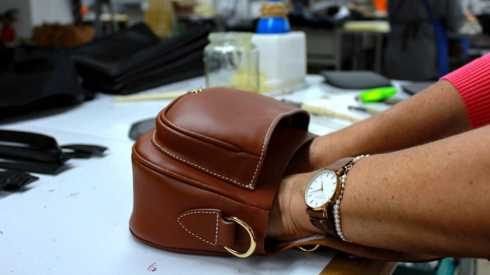 Hands finishing a tan leather crossbody bag in the workshop